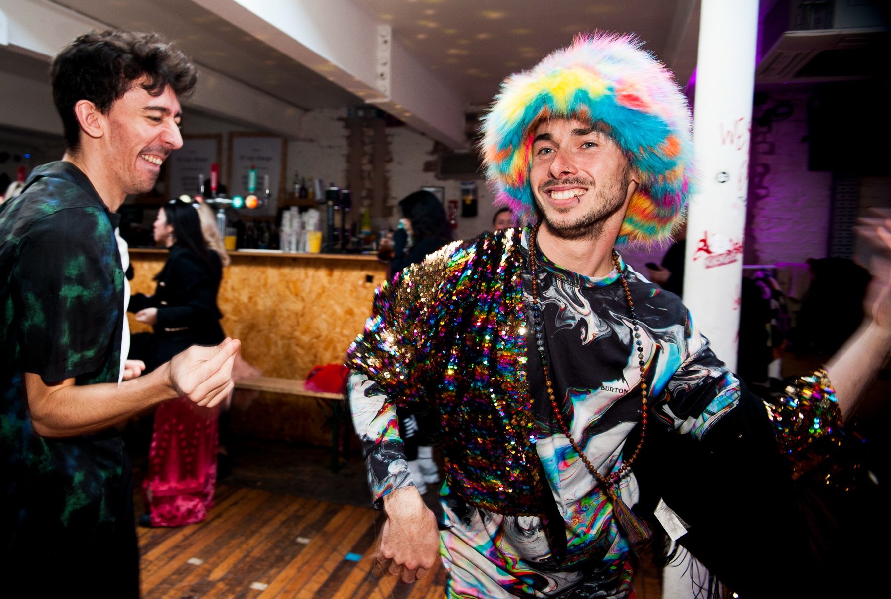 Two smiling dancers, one wearing a colourful furry hat and sequins